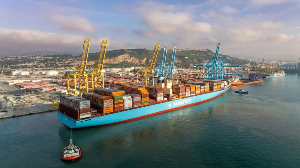 Buque portacontenedores Evelyn Maersk atracado en APM Terminals, rodeado de grúas portuarias y contenedores, con remolcador al frente y vista panorámica del puerto y ciudad al fondo.