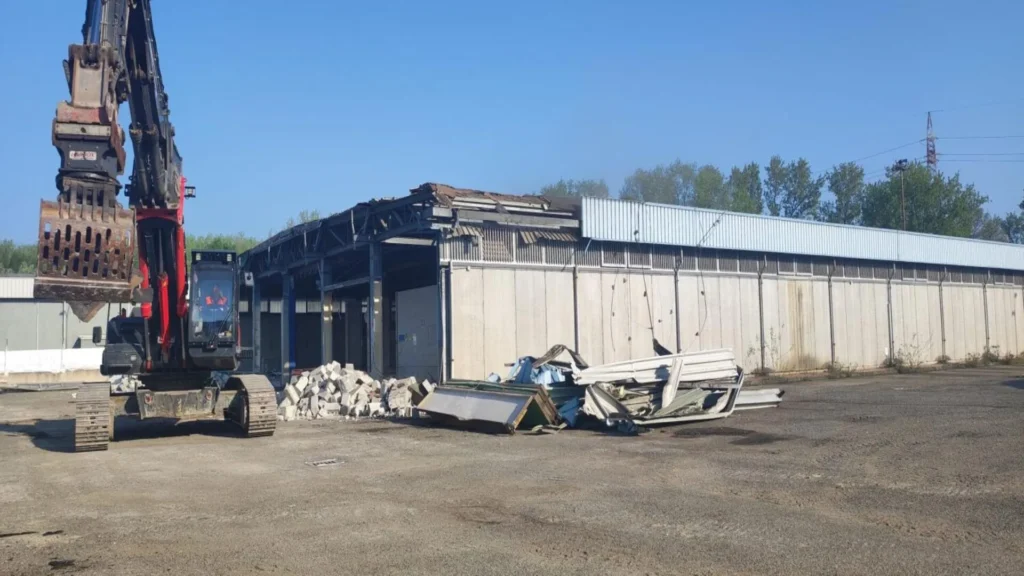 Excavadora con pinza hidráulica demoliendo parte de un almacén industrial de concreto y acero, con escombros en el suelo