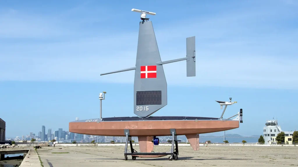 USV de Saildrone estacionado en tierra con la bandera de Dinamarca visible en su vela, y la ciudad de fondo.