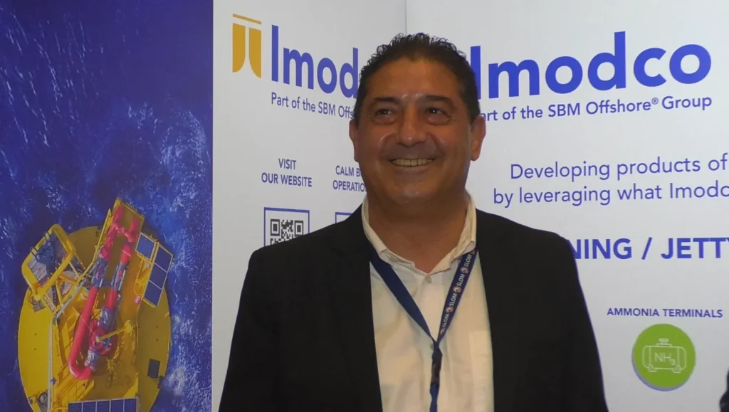 Ludovic Poix, sonríe frente a un stand de la compañía durante el evento SLOM 2025, con gráficos de boyas CALM y logotipos corporativos en el fondo.