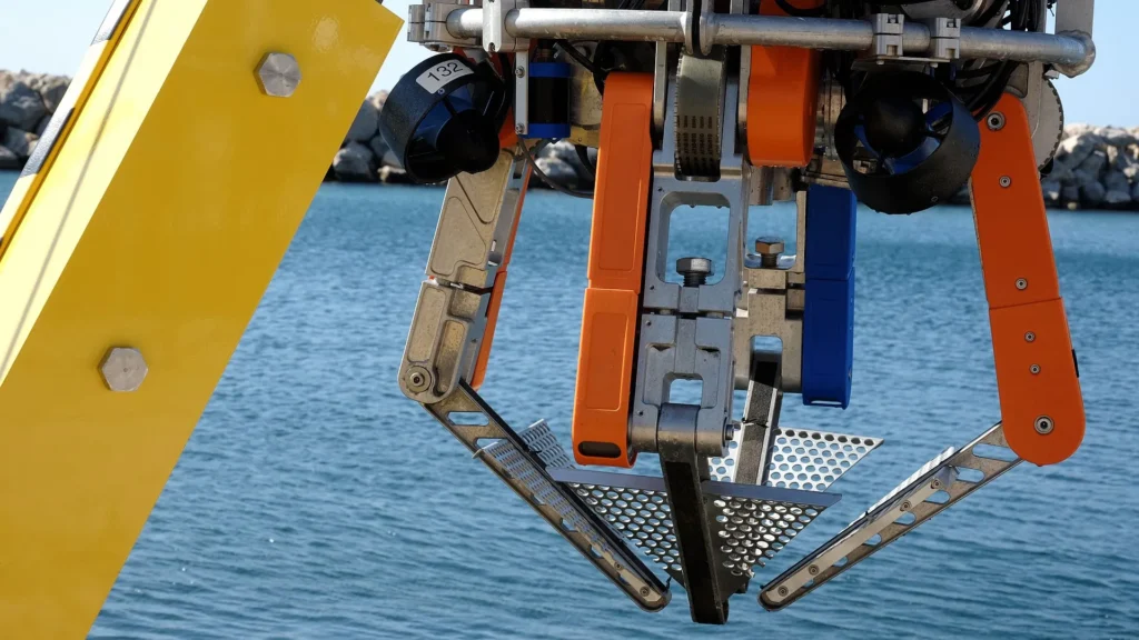 Robot submarino autónomo con estructura metálica y componentes en naranja y azul, suspendido sobre el agua antes de iniciar operaciones de recolección de basura marina.