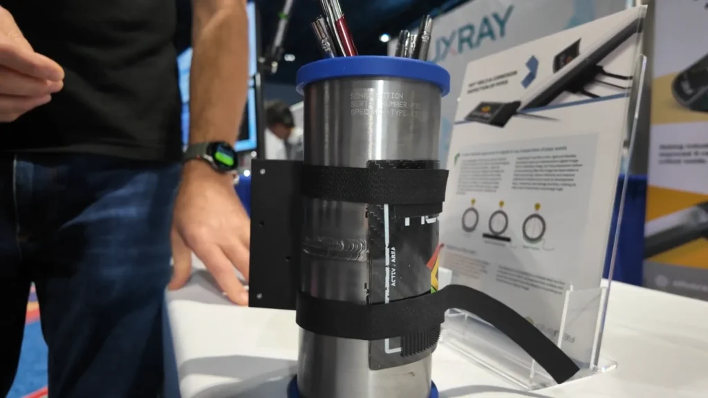A stainless steel cylindrical device with black Velcro straps holding a Silveray digital X-ray detector, displayed on an exhibition table during the ASNT 2025 convention, as an example of innovation in digitization of welds.